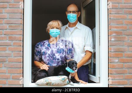 Porträt eines älteren Paares mit Hund aus dem Fenster schauen, tragen chirurgische Masken - alte Menschen Schutz vor COVID-19, Quarantäne, Pflegeheim und h Stockfoto