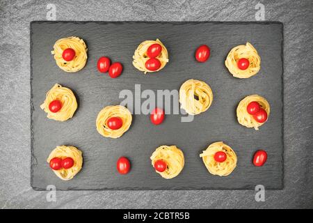 Nahaufnahme von Nestern von Tagliatelle Nudeln mit Ei-förmigen Mini roma Tomaten auf einem rustikalen Holztisch mit Sackleinen, Stockfoto