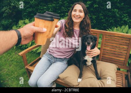 POV Bild des jungen Paares Toasting mit Bambus-Kaffeetassen sitzen im Sommergarten mit Hund - Konzept der glücklichen Familie, nachhaltiges Leben, Kunststoff re Stockfoto