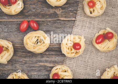 Nester von Tagliatelle Nudeln mit Ei-förmigen Mini roma Tomaten auf einem rustikalen Holztisch mit Sackleinen, Stockfoto