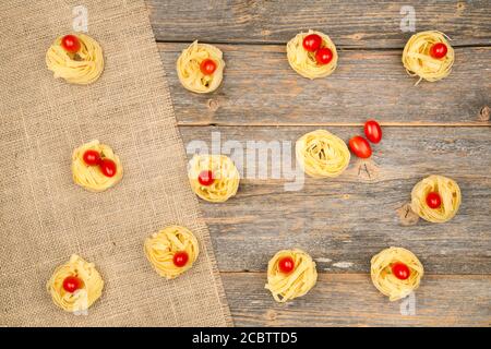 Nahaufnahme von Nestern von Tagliatelle Nudeln mit Ei-förmigen Mini roma Tomaten auf einem rustikalen Holztisch mit Sackleinen, Stockfoto