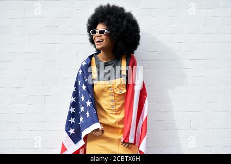 Glückliche junge afrikanische Frau eingewickelt in USA Flagge steht gegen weiße Wand. Stockfoto