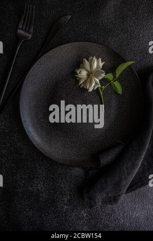 Minimalistisches Tischdekor mit schwarzem Steinzeug und Besteck mit weißer Rosenblüte auf schwarzem Steintisch mit Kopierfläche Stockfoto