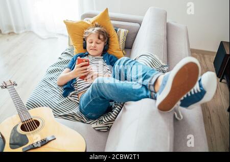 Pre teen Junge mit Gitarre auf gemütlichen Sofa gekleidet Casual Jeans und neue Sneakers Musik hören und chatten mit drahtlosen Kopfhörern verbunden w Stockfoto