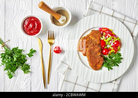 Panierte Lammkoteletts serviert mit Tomaten, roten Zwiebeln, grünem Pfeffer-Salat auf einem weißen Teller mit goldener Gabel und Messer auf einem Holztisch, flach legen Stockfoto