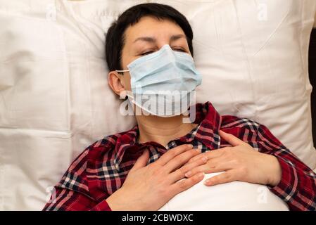 Kranke junge Frau in medizinischer Maske im Bett liegend Gefühl Brustschmerzen und keuchend. Kurzhaarige Brünette mit Symptomen von covid-19 oder Lungenentzündung zu Hause Stockfoto