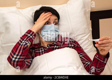 Kranke Frau im Bett in medizinischer Maske mit Fieber Blick auf Thermometer mit Angst und Überraschung. Mittlere Alter kurzhaarige Brünette auf selbst-Isolation Stockfoto