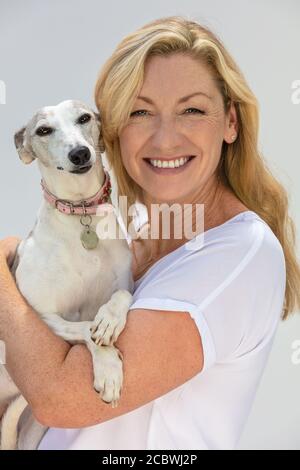 Porträt einer attraktiven mittleren Alters blonde Frau lächelnd mit Perfekte Zähne halten ihr Haustier Peitschenhund Stockfoto