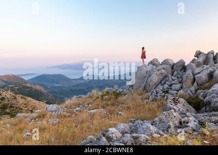 Eine Frau steht auf einem Felsvorsprung in den Bergen und blickt auf die Küste, Lefkada, Ionische Inseln, Griechenland Stockfoto