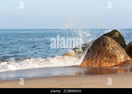 An einem sonnigen Tag trifft eine schöne Welle auf die Felsen an einem Sandstrand. Stockfoto