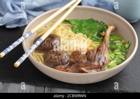 Chinesische Entennudelsuppe mit Shiitake-Pilzen, Spinat und Eiernudeln in einer braunen Schüssel mit Essstäbchen Stockfoto