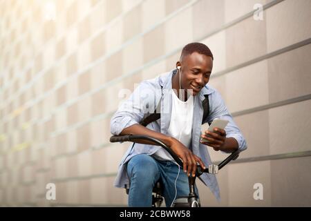 Lächelnder afroamerikanischer Kerl mit Fahrrad Musik vom Handy in der Nähe von Ziegelsteinmauer hören, freier Platz Stockfoto