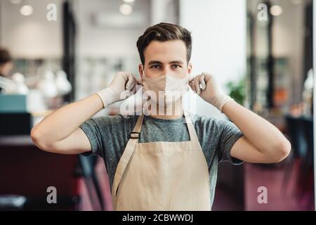 Junger Mann Barbier setzen auf Schutzmaske am Friseurladen Stockfoto