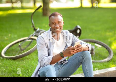 Lächelnder schwarzer Kerl mit Handy, der in der Nähe seines Fahrrads sitzt Im Park Stockfoto