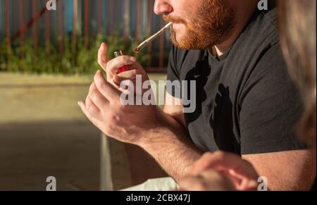 Junger Mann mit rotem Bart raucht Weed Joint auf der Straße bei Sonnenuntergang. Stockfoto