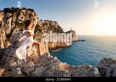 Ein Mädchen in weißem Kleid blickt auf den Leuchtturm an der Spitze des Kap Lefkada bei Sonnenuntergang, Lefkada, Ionische Inseln, Griechenland Stockfoto