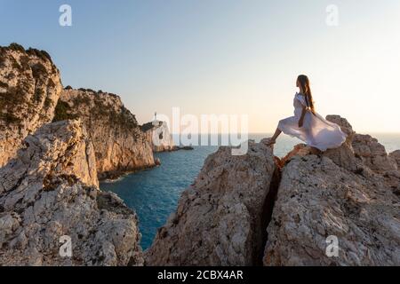 Ein Mädchen in weißem Kleid blickt auf den Leuchtturm an der Spitze des Kap Lefkada bei Sonnenuntergang, Lefkada, Ionische Inseln, Griechenland Stockfoto