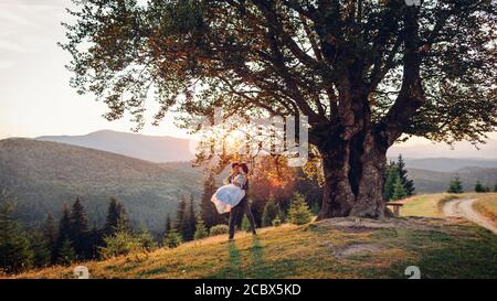 Liebevolles Brautpaar umarmt sich in den Bergen bei Sonnenuntergang. Bräutigam hält Braut auf Händen. Hochzeit im Sommer Karpaten. Stockfoto