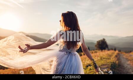 Schöne Braut tanzen in blauen Hochzeitskleid in den Bergen bei Sonnenuntergang. Frau wirft Schleier hält Blumen. Stockfoto