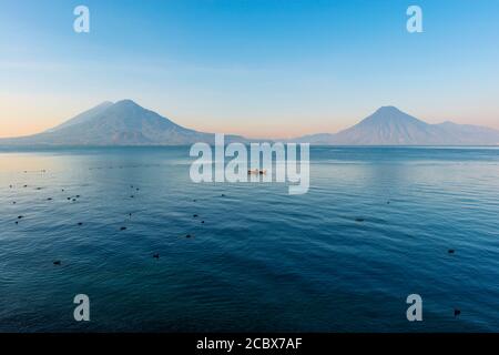 Der Vulkan San Pedro, Toliman und Atitlan bei Sonnenaufgang mit einem Fischer in Fischerboot und Blässhühner (Fulica atra), Atitlan See, Panajachel, Guatemala. Stockfoto