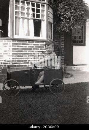 Um die 1940er Jahre, historischer, zwitschernder kleiner Junge, der in seinem Metall-Pedalauto sitzt, handgefertigt, mit Kastenkonstruktion, Drahträdern und kleiner Tür, England, Großbritannien. Stockfoto