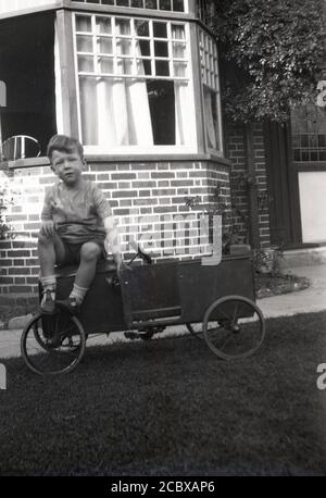 Um die 1940er Jahre, historischer, zwitschernder kleiner Junge, der auf der Motorhaube seines Metall-Pedalautos sitzt, handgefertigt, aufgrund seiner Kastenkonstruktion, Drahträder und kleinen Tür, England, Großbritannien. Stockfoto