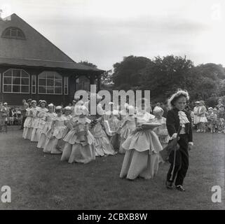 1950er Jahre, historisch, ein kleiner Junge in einem Regency-Kostüm mit Spitzenkräften, die die „Rose Queen“ und ihre Gefolgschaft junger Mädchen führen, während sie durch den lokalen Park, Farnworth, Lancashire, England, Großbritannien, spazieren. Seit den 1880er Jahren findet das jährliche Rose Queen Festival traditionell im Juni statt, das nach den Kriegen der Rosen (1455–87) in vielen Städten und Dörfern Großbritanniens, insbesondere im County Lancashire, bekannt als Red Rose County, jährlich stattfindet. Stockfoto