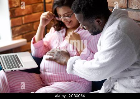 Schwarzer Mann ist freundlich zu seiner schwangeren kaukasischen Frau, junge männliche Schlaganfall Bauch, Frau im Pyjama sitzt mit Laptop zu Hause, verliebt Stockfoto