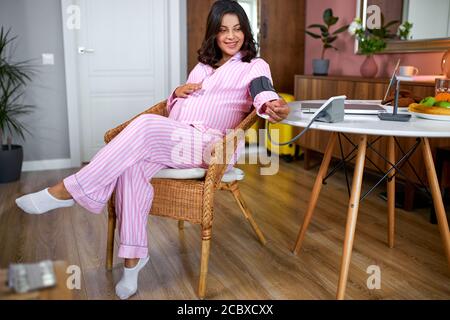 kaukasische schwangere Frau misst den Blutdruck mit automatischem Blutdruckmessgerät zu Hause, sitzen hinter dem Tisch in Pyjamas, am Morgen, allein Stockfoto
