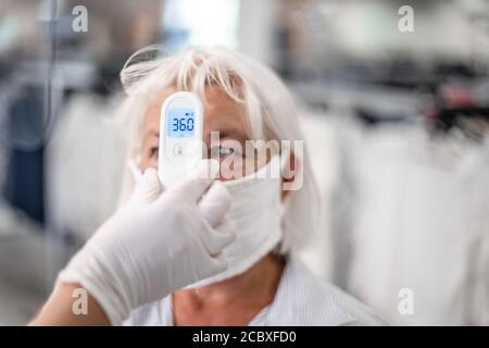 Hand hält Infrarot-Thermometer, um die Temperatur auf verschwommene Frau mit Gesichtsmaske auf dem Hintergrund zu messen. Stockfoto