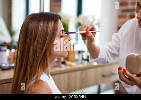 Prozess der Herstellung Make-up in Schönheitssalon. Visagiste arbeiten mit Pinsel auf Modell Gesicht. Anwendung Ton auf die Haut. Professionelle Make-up Künstler malt Modell Stockfoto