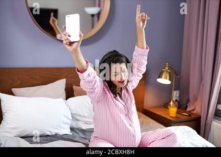 Schwangere Frau singt ihre Lieblingslieder mit jubelnder Musik, während sie morgens zu Hause auf dem Bett sitzt, entzückende Frau mit Kopfhörern genießt die Erwartung Stockfoto