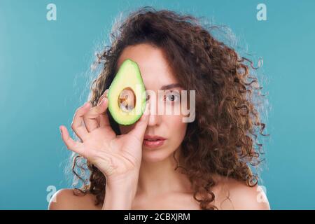 Junge Frau, die Sie mit dem linken Auge anschaut, während sie hält Avocado von einem anderen Stockfoto