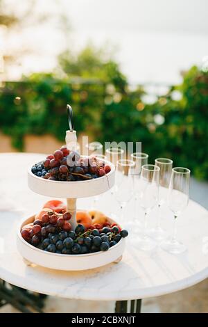Zweistufiges Lebensmittelregal mit Trauben und Früchten auf einem weißen Tisch mit leeren Champagnergläsern. Stockfoto