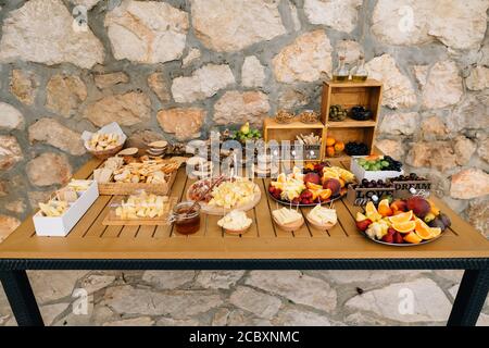 Holztisch serviert mit Köstlichkeiten aus Käse, Obst und Schinken vor dem Hintergrund einer Steinmauer. Stockfoto