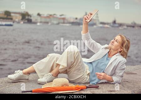 Schöne aktive ältere weiße Frau über 62 Jahre alt mit Blonde Haare und in lässigen und eleganten Tuch sitzt In der Nähe des Flusses in Sankt-Petersburg mit Stockfoto