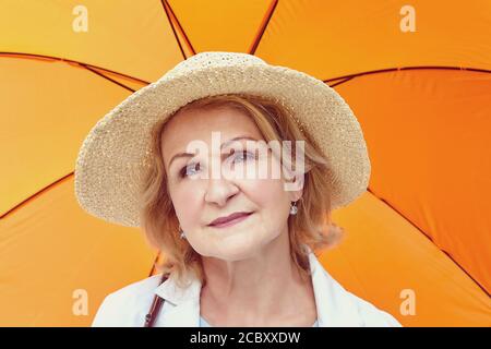Schöne weiße reife Frau etwa 62 Jahre alt mit blonden Haaren in Hut und unter orangenen Regenschirm ist zu Fuß in den öffentlichen Park. Alter Kaukasier hübsch Stockfoto