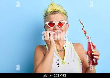 Attraktive reife Frau mit Fingern im Mund hält eine Flasche Saft. Close up Porträt, isoliert blauen Hintergrund, Studio erschossen Stockfoto