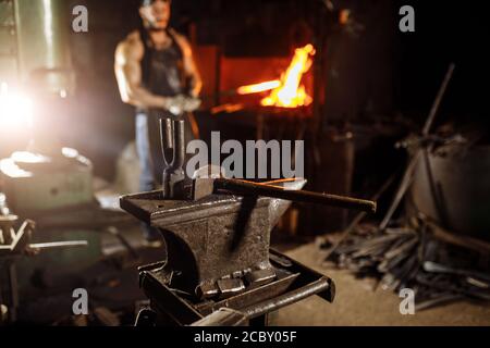 Nahaufnahme von Hammer und anderen Instrumenten, Schmiedeausrüstung, Schmiedekunst. Muskulöser junger Mann in der Nähe von Ofen im Hintergrund Stockfoto