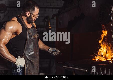 Junger kaukasischer Mann Schmied, starker Eisenarbeiter in der Werkstatt, Arbeitsplatz, isolierter dunkler Raum mit Ofen Stockfoto