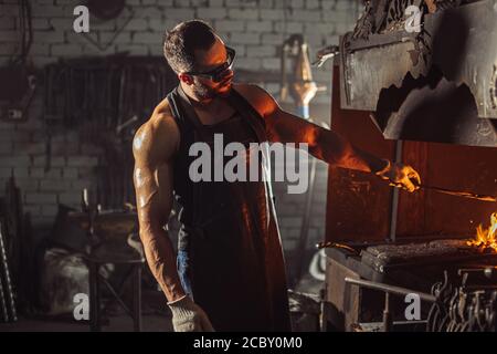 Muskulöser Erwachsener mit Bart trägt Lederschürze in der Werkstatt, Heizung. Gehärtetes Metall im Ofen Stockfoto