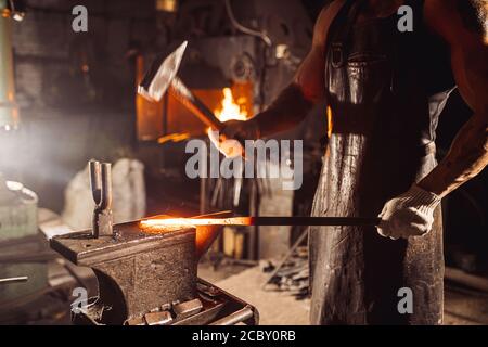 Muskulöser Erwachsener mit Bart trägt Lederschürze in der Werkstatt, Heizung. Gehärtetes Metall im Ofen Stockfoto