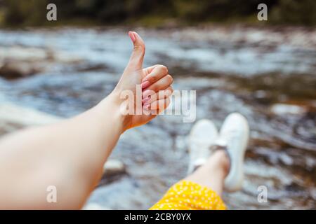 Frau entspannt am Fluss des Berges zeigt Daumen nach oben genießen Landschaft. Reisender sitzt auf Felsen. Sommerurlaub Stockfoto