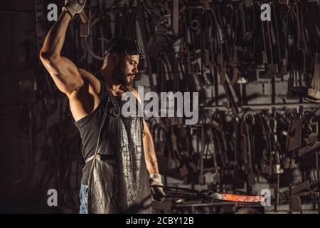 Starke muskulöse brutal zuversichtlich Schmied Mann Formen roten heißen Metall mit Hammer isoliert in der Werkstatt, trägt Lederschürze, dunklen Raum Stockfoto