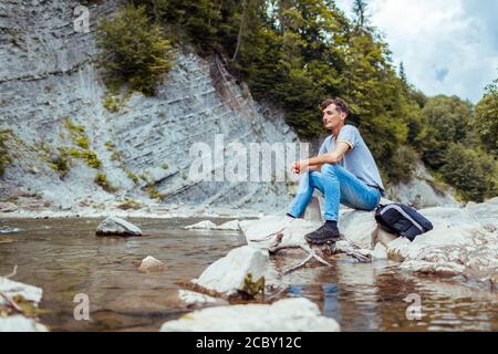 Junger Mann, der sich am Bergfluss entspannt und die natürliche Landschaft genießt. Reisender Rucksacktourist sitzt auf Felsen. Sommerurlaub Stockfoto