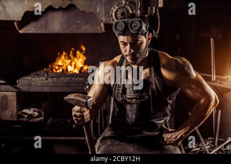 Portrait von nachdenklichen muskulösen Schmied hält Hammer und Blick nach unten, trägt Lederuniform. Ofen im Hintergrund Stockfoto