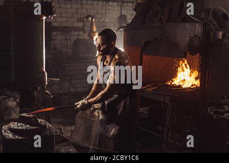 Porträt des Schmieds in Leder Schürze hält heißes Metall, trägt braune Leder Schürze, Raum mit Ofen, Feuer Stockfoto
