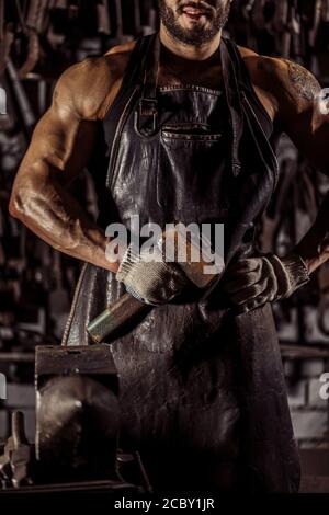 Unerkennbarer Mann mit muskulösem Körper, junger Schmied hält Hammer in den Händen, haben Bart. Instrumente im Hintergrund Stockfoto