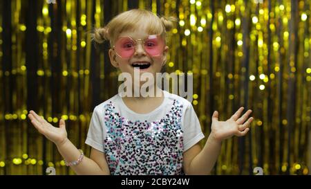 Kind tanzt, zeigt Erstaunen, täuscht herum, lächelt, schaut überrascht schockiert. Wenig Spaß blonde Kind Teenager Teenager Mädchen 4-5 Jahre alt in Sonnenbrille posiert isoliert auf Hintergrund mit Folie Vorhang Stockfoto