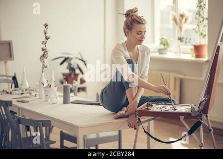 Talentierte Künstlerin sitzt auf dem Tisch und Zeichnung ihr Bild zu Hause. Volle Länge Seitenansicht Foto. Interesse, Hobby-Konzepte Stockfoto